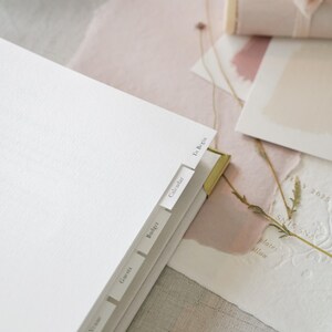 May include: A white planner with labeled tabs, including "To Begin," "Calendar," and "Budget." A pencil and a small brass ruler are in the foreground. Soft pink paper and a rolled scroll tied with a ribbon are in the background. The scene has a neutral, elegant aesthetic.