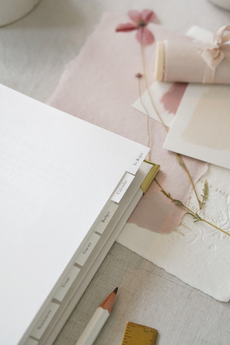 May include: A white planner with labeled tabs, including "To Begin," "Calendar," and "Budget." A pencil and a small brass ruler are in the foreground. Soft pink paper and a rolled scroll tied with a ribbon are in the background. The scene has a neutral, elegant aesthetic.