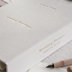 May include: A light gray wedding planner with gold lettering that reads "Wedding Planner" and "Gable & Grain." A pink pen and a gold paper clip are next to the planner. The planner has gold corner protectors.