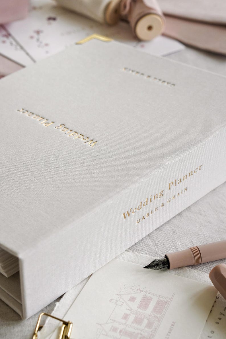 May include: A light gray wedding planner with gold lettering that reads "Wedding Planner" and "Gable & Grain." A pink pen and a gold paper clip are next to the planner. The planner has gold corner protectors.
