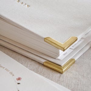 May include: Close-up of a stack of off-white linen-covered books with gold-colored metal corner protectors. The top book has gold lettering that reads "LISA & GRAIN". A "Save our date" card with floral accents is visible.