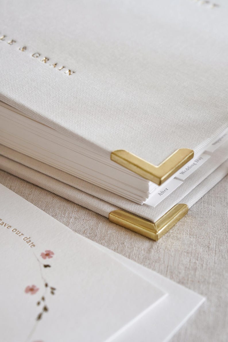 May include: Close-up of a stack of off-white linen-covered books with gold-colored metal corner protectors. The top book has gold lettering that reads "LISA & GRAIN". A "Save our date" card with floral accents is visible.