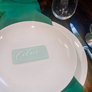 May include: A white plate with a teal green place card that says "Colin" on it. The plate is on top of a green napkin.