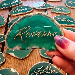 May include: Green agate place cards with gold lettering. The name "Roxanne" is featured prominently on one card, held by a hand with a colorful manicure. Other cards with names like "Olivia" and "Jillian" are visible. The cards have gold edges.