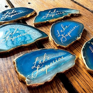 May include: Six blue and white agate coasters with gold edges are arranged on a wooden surface. Each coaster has the words "Lake Superior" written in elegant script. The coasters vary in shape and size.