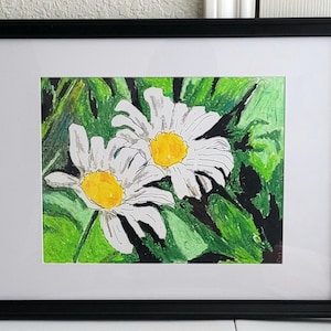 May include: Framed watercolor painting of two daisies with white petals and yellow centers, set against a backdrop of green foliage. The artwork is framed in a black frame with a white mat.