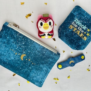 May include: A collection of celestial-themed accessories, including a blue zippered pouch with a crescent moon and star design, a small coin purse with "Reach for the Stars" embroidery, a key fob, and an owl keychain. The items are on a white surface.