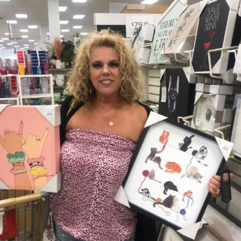 May include: A woman holding two framed art pieces in a store. One is a pink canvas with hand gestures and bracelets. The other is a black framed print with cat illustrations. Shelves with other art pieces are in the background.