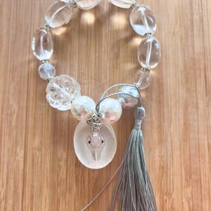 May include: A clear beaded bracelet with oval and round beads. The bracelet has a decorative pendant with a bird design, pearl beads, and a gray tassel. The bracelet is on a wooden surface.