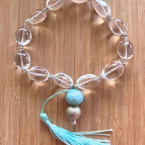 May include: Clear oval beads bracelet with silver accents. A light blue and silver bead cluster hangs from the bracelet, with a matching light blue tassel. The bracelet is displayed on a wooden surface.