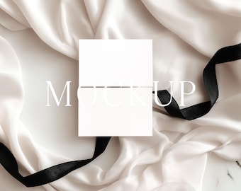 May include: A white rectangular card with the word "MOCKUP" printed on it, resting on a backdrop of draped, off-white fabric. A black satin ribbon is draped across the scene, adding a contrasting element.