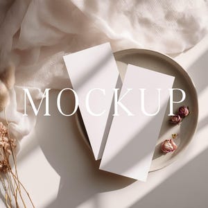May include: Two blank white rectangular paper cards positioned on a round ceramic plate. The scene includes dried beige flowers and a soft white fabric draped nearby. The word MOCKUP is printed in large white capital letters across the center.