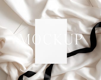May include: A blank white card with the word "MOCKUP" in a light font is centered on a background of ivory fabric and a black ribbon. The fabric is draped and folded, creating soft shadows and textures.