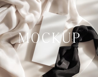 May include: A white rectangular card with the word "MOCKUP" in the center, resting on a backdrop of ivory fabric and a black silk ribbon. The scene is lit with soft, natural light, creating shadows.
