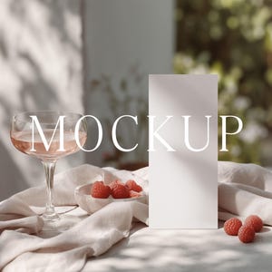 May include: A champagne coupe glass with a pink drink, a small bowl of raspberries, and a blank white card on a linen cloth. The word "MOCKUP" is displayed across the image.