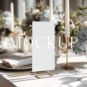 May include: A table setting with a white rectangular sign in a gold stand, with the word "MOCKUP" in white. The table is set with plates, silverware, candles, and floral arrangements. The overall theme is elegant and sophisticated.