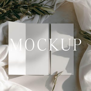 May include: A tri-fold brochure mockup featuring a clean, light gray paper surface with sharp, defined creases. The word MOCKUP is printed in large, white, serif capital letters across the center of the paper panels, which are displayed flat.