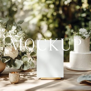 May include: A blank white rectangular sign on a gold stand, positioned on a wooden table next to a large floral arrangement of white roses and eucalyptus, a two-tiered white cake, a small textured candle holder, and a glass of champagne.