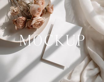 May include: A mockup scene featuring a white plate with dried roses, two blank cards, and a draped fabric. The word "MOCKUP" is printed on the image. The scene is lit with natural light, creating shadows.