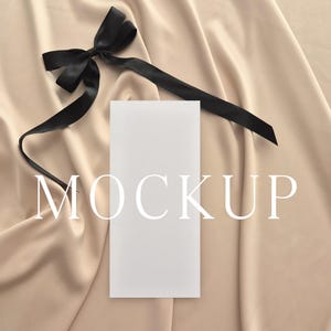 May include: A white rectangular card with the word "MOCKUP" in a serif font, placed on a beige satin fabric. A black satin ribbon is tied in a bow and placed near the card.