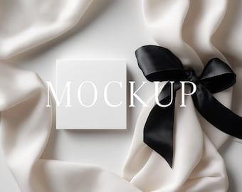 May include: A white square box with the word "MOCKUP" printed on it, next to a black satin bow. The box and bow are set against a backdrop of flowing white fabric.
