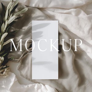 May include: A white rectangular mockup card with the word "MOCKUP" in elegant font, resting on a textured, neutral-toned fabric. Olive branches and dried flowers are arranged to the left, casting shadows.