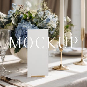 Puede incluir: Una mesa con una tarjeta rectangular blanca con la palabra "MOCKUP" en blanco, un arreglo floral de flores azules y blancas, dos copas de vino y candelabros dorados.