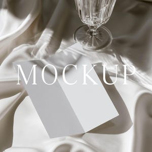 May include: A blank white folded card mockup positioned on draped cream-colored satin fabric next to a clear glass goblet. The word MOCKUP is printed in large white capital letters across the center of the card, which features a clean surface.