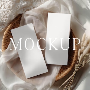May include: Two blank, rectangular white paper cards positioned vertically on a round woven wicker tray. The tray is lined with a soft, draped off-white fabric and accompanied by a small bundle of dried beige grass stems on the side.