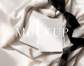 May include: A white card and envelope mockup with the word "MOCKUP" in the center. The stationery is set on a cream-colored fabric with a black ribbon. The scene is lit with soft, natural light.