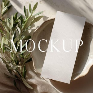 May include: A mockup scene featuring a white rectangular card resting on a ceramic plate with a textured rim. Olive branches with green leaves are placed on a beige cloth. The word "MOCKUP" is printed on the image.