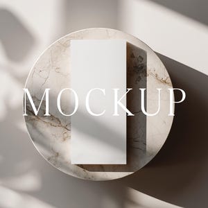 May include: A white rectangular card sits atop a round marble surface, with the word "MOCKUP" in white text. The marble has a beige and brown pattern. Shadows from a nearby plant create a natural aesthetic.