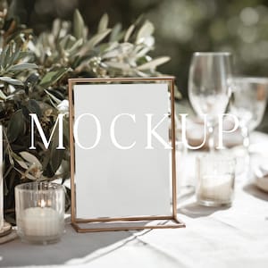 May include: A table setting with a rectangular gold frame with the word "MOCKUP" in white. A lit candle, glass candle holders, and wine glasses are on the table. Olive branches are in the background.