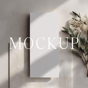 May include: A white rectangular mockup with the word "MOCKUP" in white text. The mockup is positioned on a textured, light-colored surface, with a sprig of greenery and sunlight casting shadows.