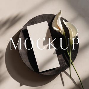 May include: A mockup scene featuring a dark gray circular tray with a white rectangular card and two white calla lilies. The word "MOCKUP" is overlaid on the scene. The background is a light-colored surface with shadows.