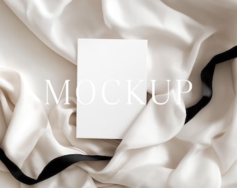 May include: A blank white card with the word "MOCKUP" in a light font is centered on a cream-colored satin fabric. A black ribbon is draped across the bottom right corner of the fabric, adding a contrasting element to the composition.