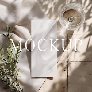 May include: A mockup scene featuring a white rectangular card, a glass of liquid, and sprigs of greenery. The word "MOCKUP" is displayed in large, elegant font. The scene is set on a textured surface with shadows.