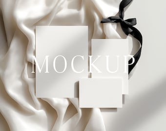 May include: A mockup featuring three white rectangular cards of varying sizes arranged on a white silk fabric. A black ribbon with a bow is draped over the cards. The word "MOCKUP" is printed on the cards.
