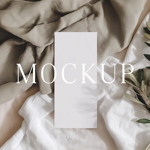 May include: A white rectangular card with the word "MOCKUP" in a serif font, placed on a bed of neutral-toned fabric. Olive branches with green leaves and small flowers are arranged on the right side.