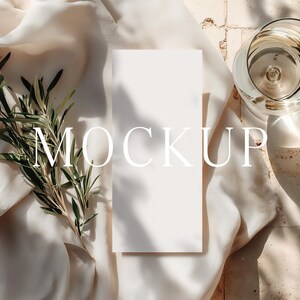 May include: A white rectangular card with the word "MOCKUP" printed on it, placed on a cream-colored fabric. A sprig of green leaves and a glass of liquid are also present, with shadows cast across the scene.