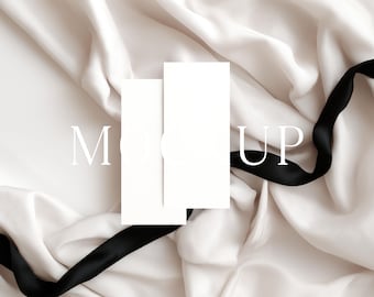 May include: A white card mockup with a circular clasp, set against a backdrop of ivory silk fabric. A black satin ribbon weaves across the scene, adding contrast. The word "MOCK UP" is printed on the image.