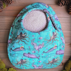May include: A teal tote bag with a circular opening and a patterned exterior. The pattern features octopuses, narwhals, and seahorses in top hats and other whimsical designs. The interior is a light gray fabric.