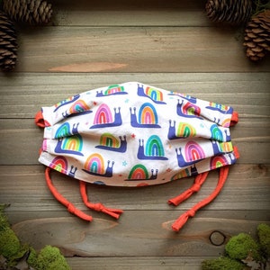 May include: White face mask with a repeating pattern of colorful rainbow snails and small stars. The mask has orange trim and orange ties for securing. The mask is made of fabric and is designed to cover the nose and mouth.