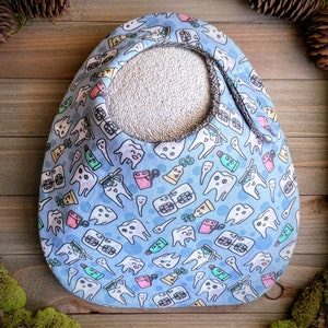 May include: A baby bib with a light blue background and a pattern of cartoon teeth, toothbrushes, and toothpaste tubes. The bib has a white terry cloth center and a gray trim. The bib is designed to catch spills and drool.