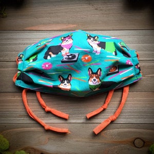 May include: A turquoise blue face mask with a colorful pattern of corgis wearing sunglasses and listening to music. The mask has orange ties.