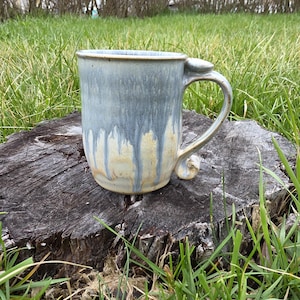Könnte beinhalten: Eine handgefertigte Keramik-Tasse mit einem einzigartigen Griffdesign. Die Tasse hat eine blau-graue Glasur mit einem Tropfeffekt, der eine beige Farbe darunter zeigt. Die Tasse steht auf einem Baumstumpf in einer grasbewachsenen Umgebung.