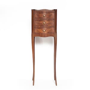 May include: A tall, narrow wooden chest of drawers featuring three stacked drawers with curved fronts and small, round brass-coloured knobs. The piece stands on four slender, elongated legs that curve outward, showcasing a rich brown wood grain.