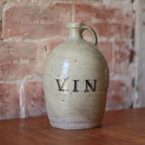 Może przedstawiać: Kremowy ceramiczny dzban z uchwytem i napisem "VIN" czarnymi literami. Dzban stoi na drewnianej powierzchni, w tle ceglana ściana.
