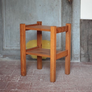Op de afbeelding: Een kleine, houten bijzettafel met twee niveaus. De tafel is gemaakt van lichtbruin hout en heeft een eenvoudig, geometrisch ontwerp. De tafel is ongeveer 61 cm hoog en 30 cm breed, met een plank in het midden.