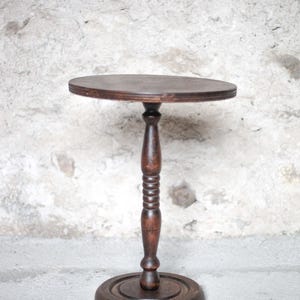 Pode incluir: Uma mesa de pedestal de madeira castanho-escura com um tampo circular, uma coluna central torneada com sulcos decorativos e uma base redonda e plana. O veio da madeira &eacute; vis&iacute;vel em toda a estrutura, exibindo um acabamento tingido profundo.